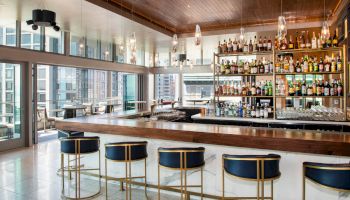 The image shows a modern bar with four chairs, a large wooden counter, and shelves of liquor bottles. There are big windows with a city view.