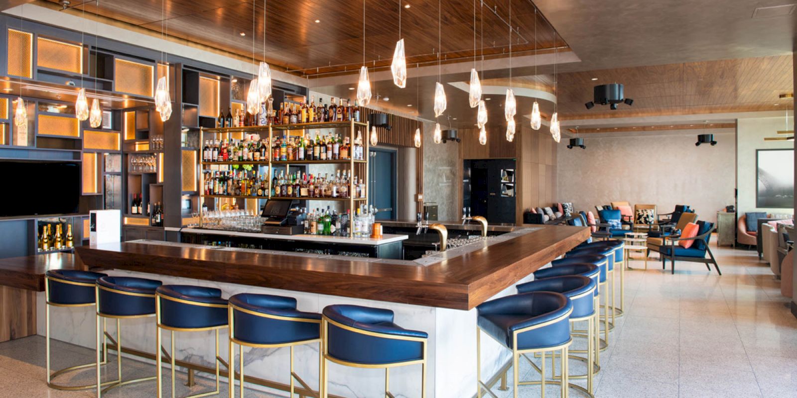 A modern bar setup with blue chairs, a wooden countertop, hanging lights, and a fully stocked drink shelf. Lounge seating is seen in the background.