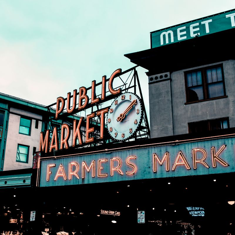 The image shows the iconic "Public Market" and "Farmers Market" signs at Pike Place Market in Seattle, Washington, with buildings and a clock.