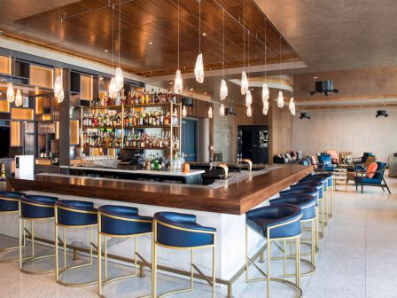 The image shows a modern bar with blue chairs, a well-stocked liquor shelf, and pendant lights. There are tables and seating areas in the background.