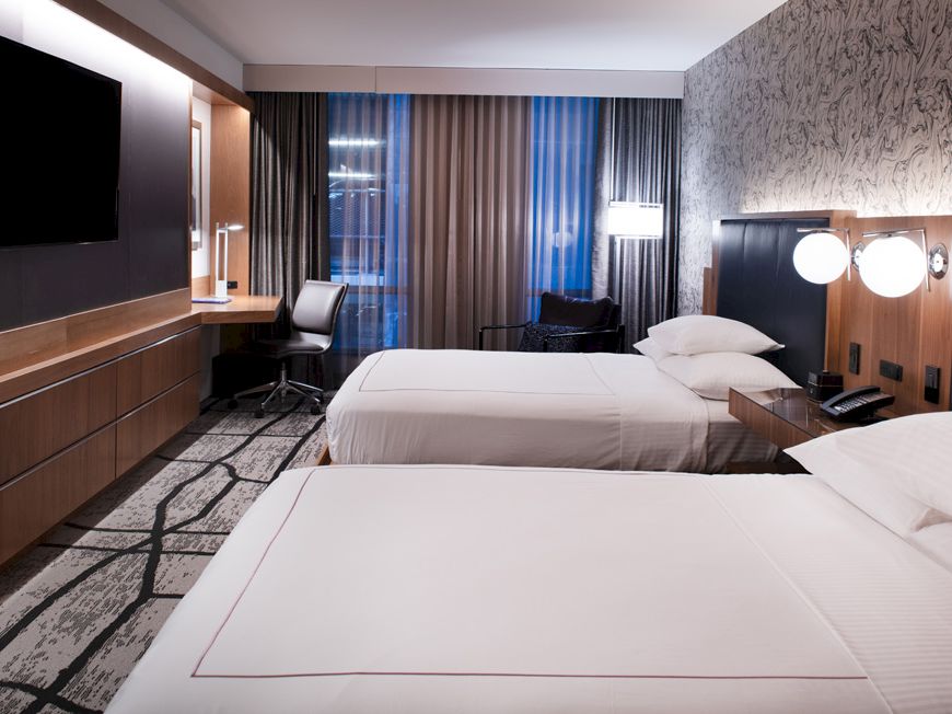 A modern hotel room features two double beds, a flat-screen TV, a desk, a chair, and ambient lighting with contemporary design elements.