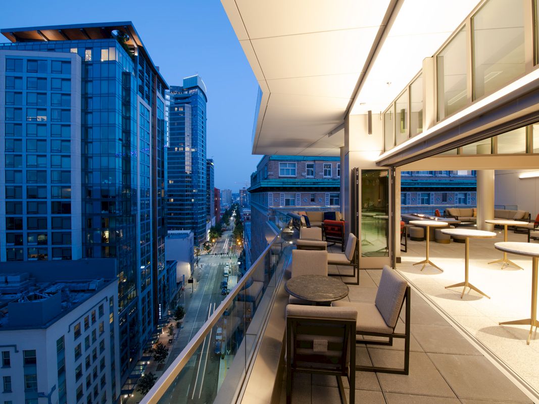 A modern balcony with seating and tables overlooks a city street lined with buildings at dusk, featuring contemporary architecture and city lights.
