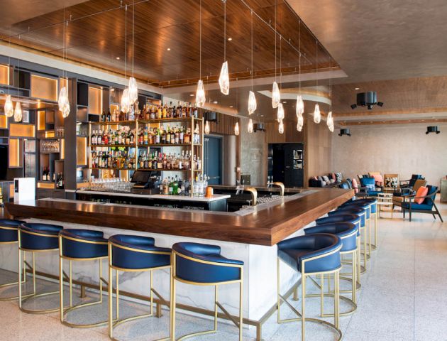 The image shows a modern, stylish bar with a long counter, blue stools, hanging lights, and a well-stocked shelf of drinks in the background.