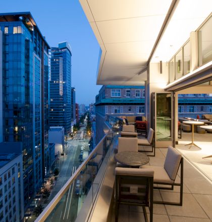 The image shows an outdoor balcony area with tables and chairs overlooking a city street, surrounded by tall, modern buildings at dusk.