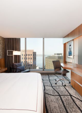 A modern hotel room with a large bed, a flat-screen TV, a desk with a chair, and a city view through floor-to-ceiling windows.