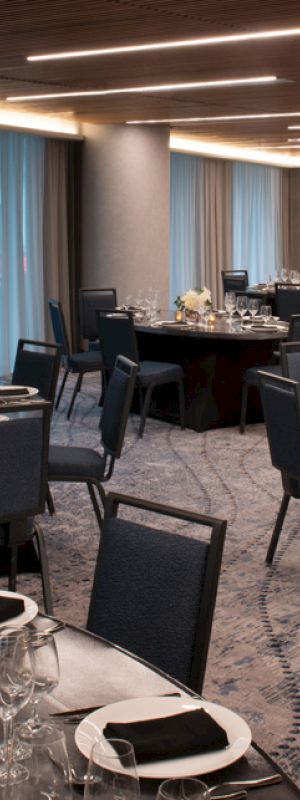 A modern, elegantly set dining room with multiple tables, black chairs, silverware, glassware, and floral arrangements, ready for a formal event.