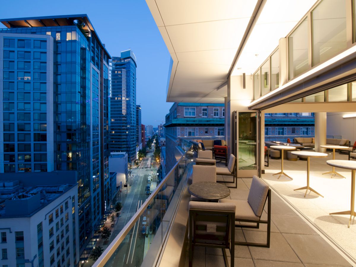A modern, glass-walled balcony with outdoor seating overlooks a city street surrounded by tall buildings at dusk.
