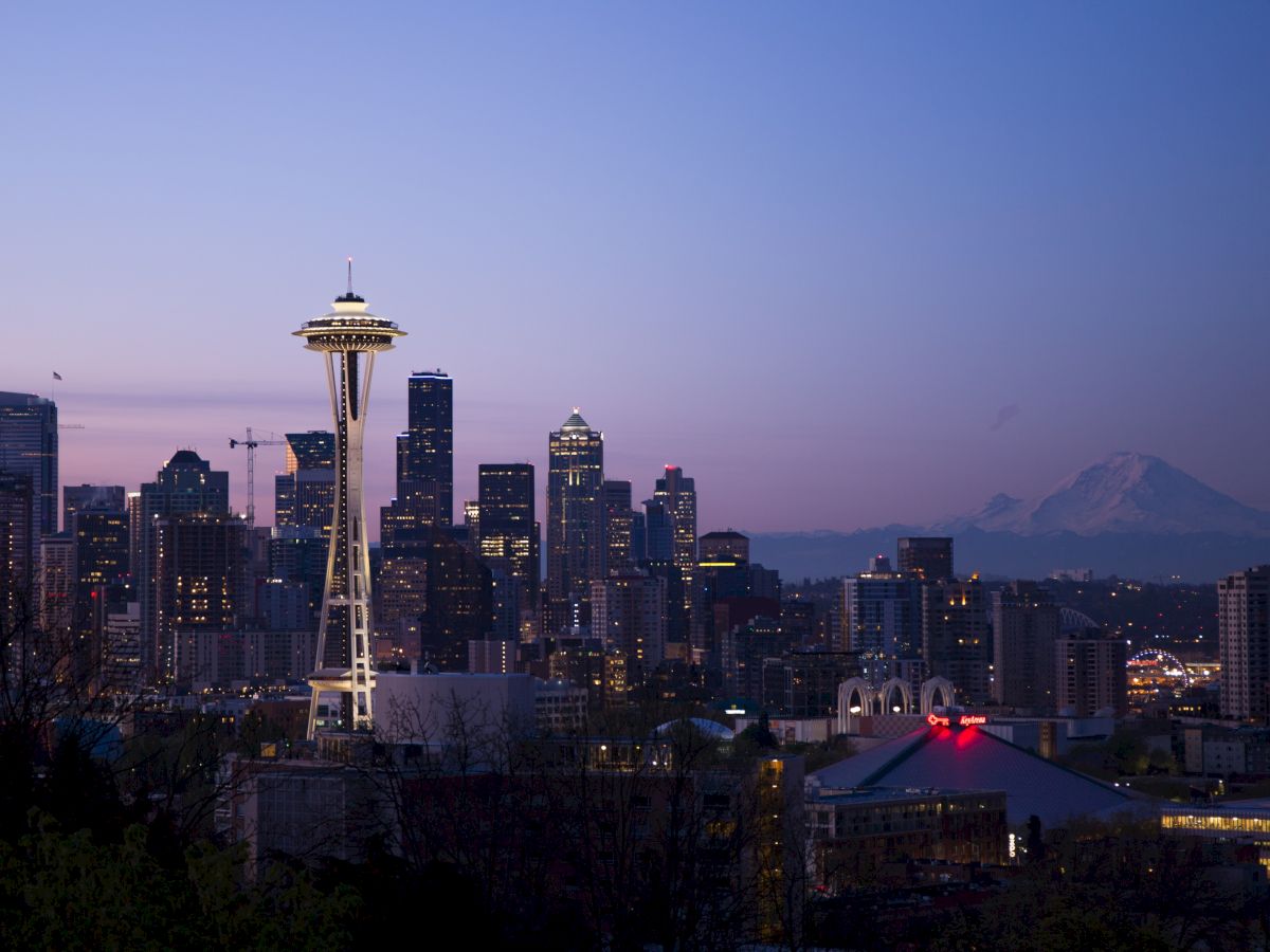 This image shows the Seattle skyline featuring the Space Needle at dusk, with Mount Rainier visible in the background against a purple and blue sky.
