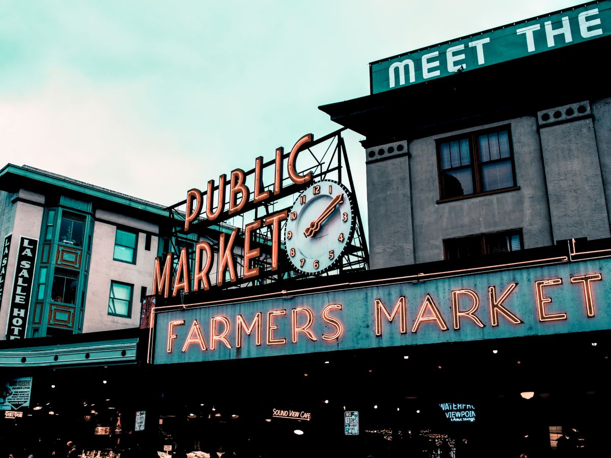 The image shows a public market with a neon "Farmers Market" sign and a clock above it, indicating a bustling community area.