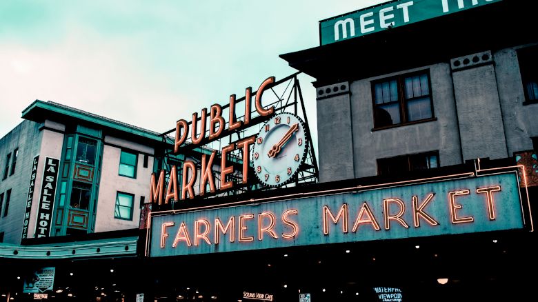 The image shows a public market with a neon "Farmers Market" sign and a clock above it, indicating a bustling community area.