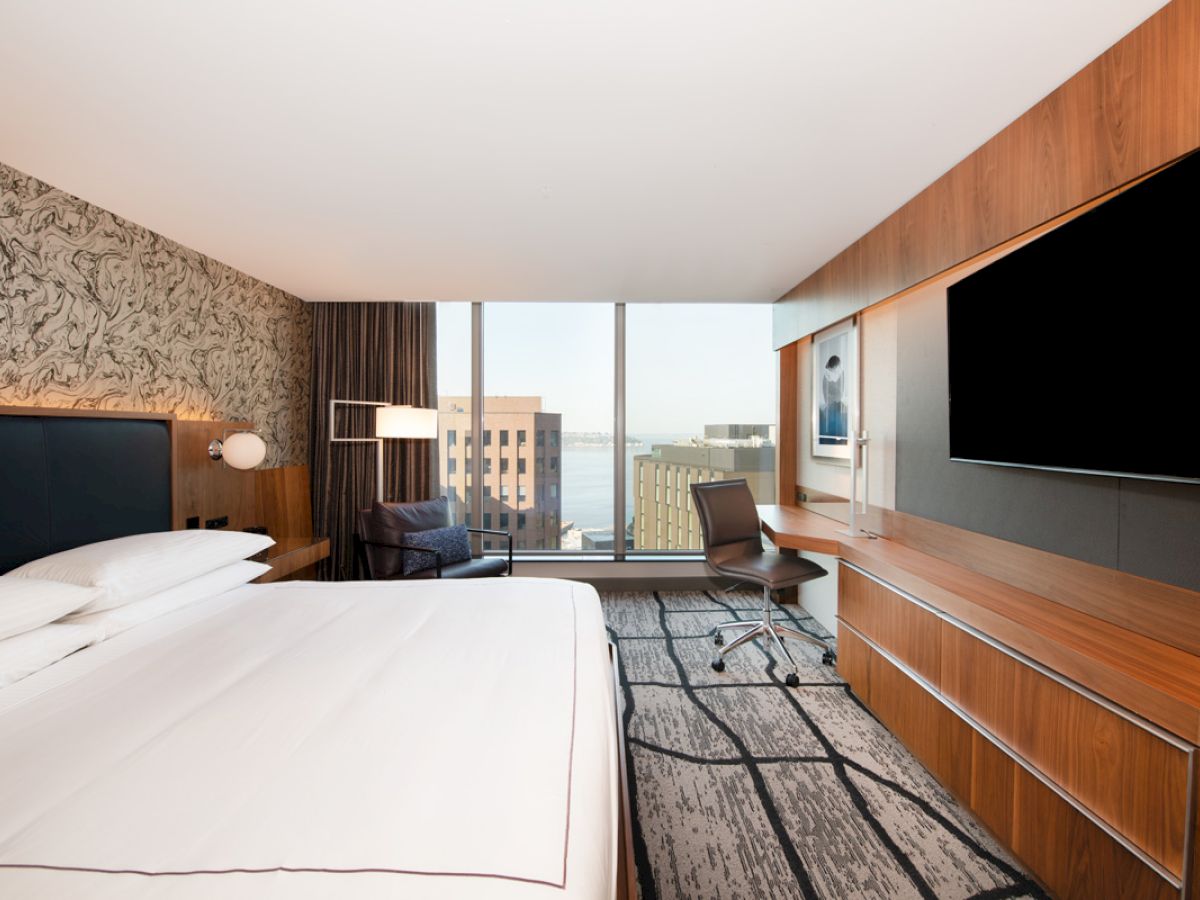 The image shows a modern hotel room with a large bed, a flat-screen TV, a desk with a chair, and a view of buildings through a large window.