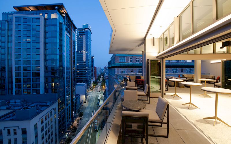 A modern urban balcony overlooks a cityscape with tall buildings at dusk. The balcony is furnished with tables and chairs.